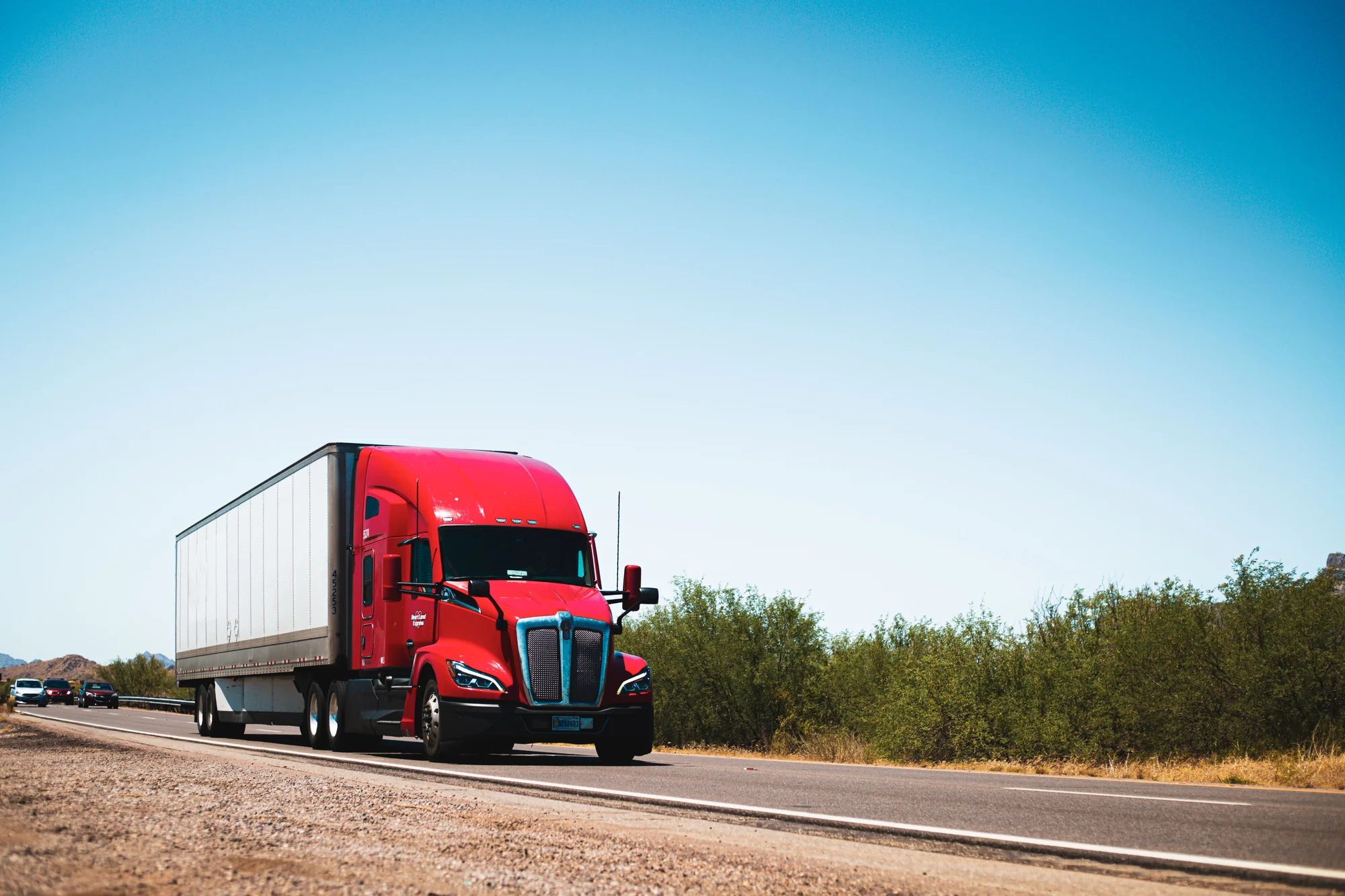 Red Kenworth semi truck pulling a white trailer along a country road